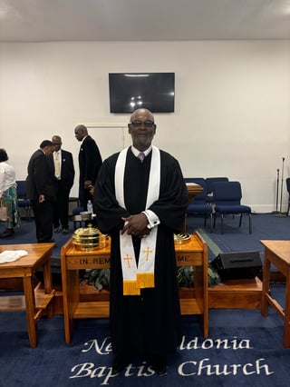 Clergy member in black robe and white stole standing in church sanctuary with other people in background