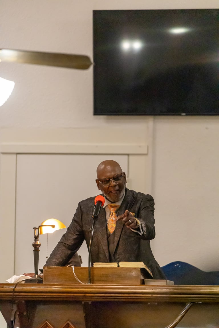 Man in glasses and dark blazer speaking at wooden podium with microphone in front of wall-mounted TV and whiteboard